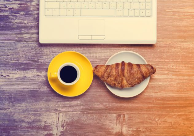 Cup of coffee and croissant near notebook on wooden table. work