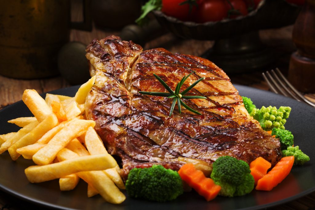 Grilled beef steak served with French fries and vegetables on a black plate.